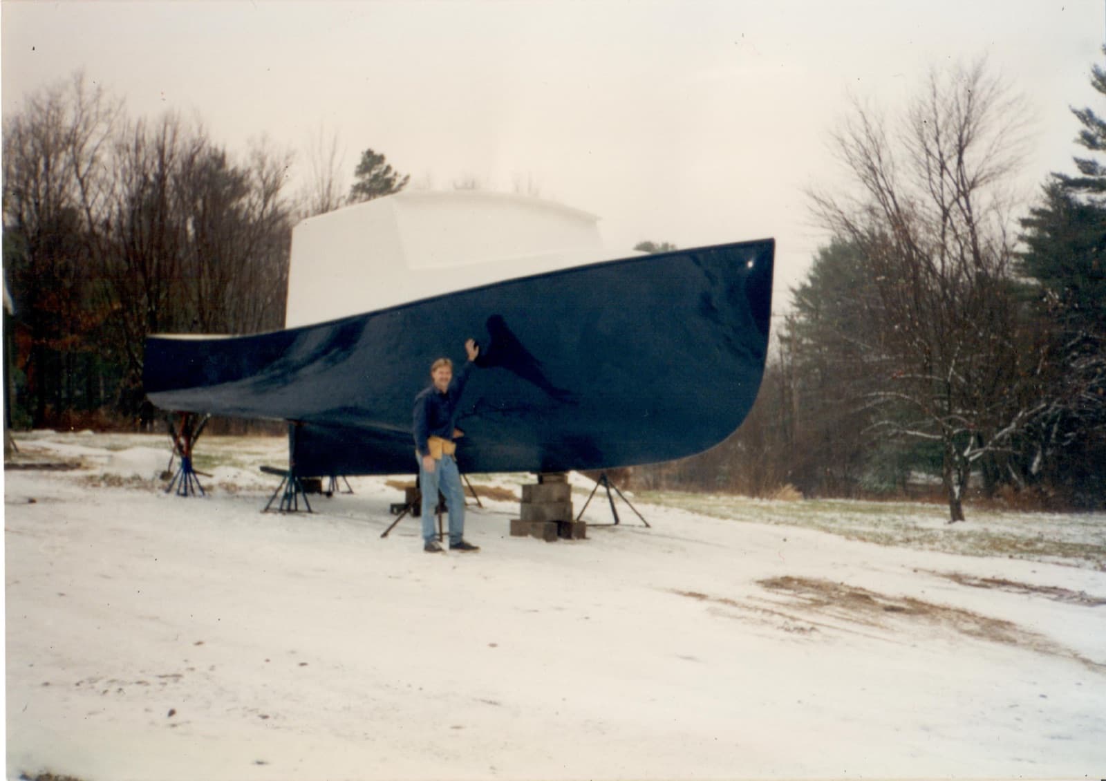 Donald Clark beside a deep-blue yacht hull on stands outside the shop in winter.
